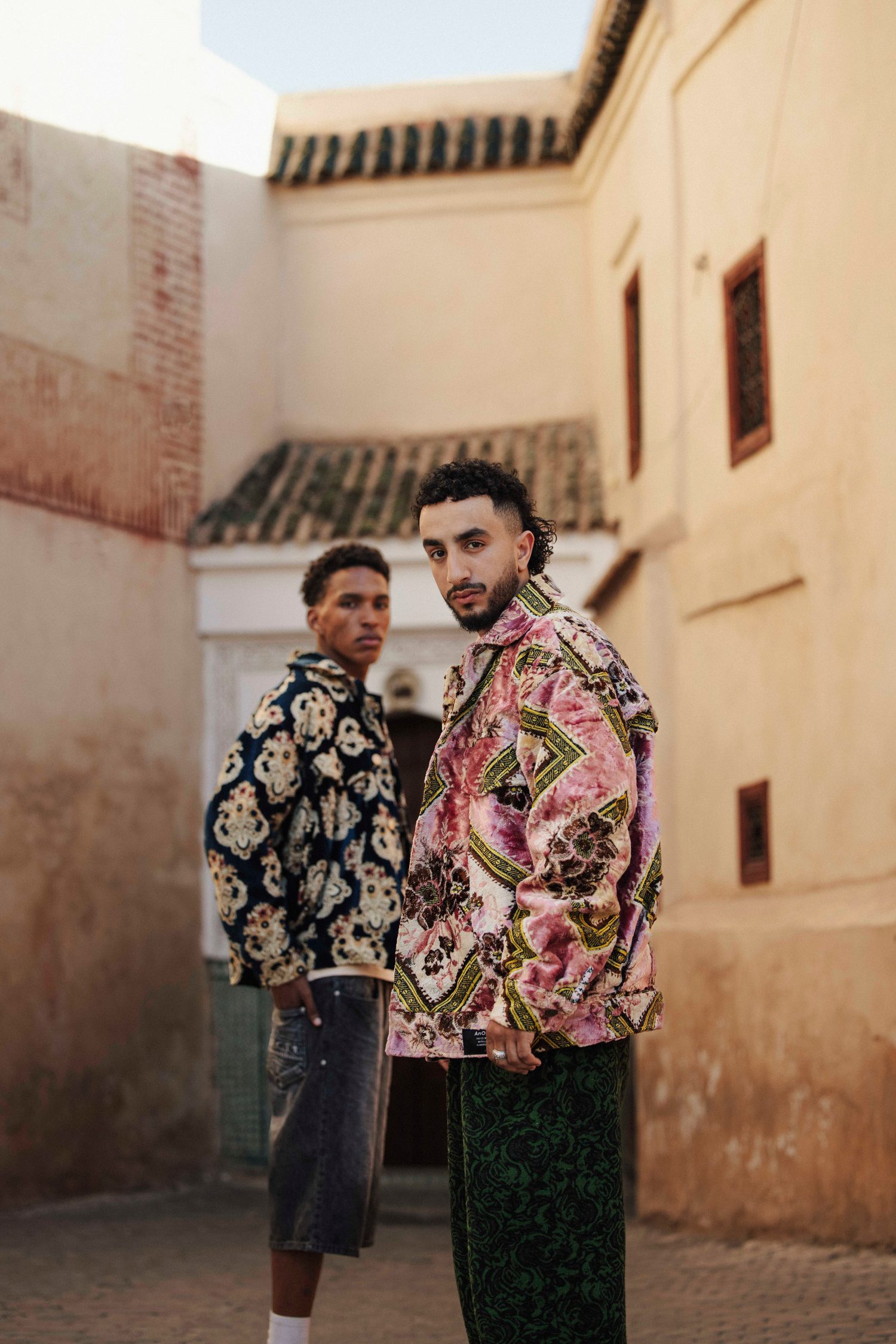 Two models in statement jackets standing in a sunlit Marrakech alley
