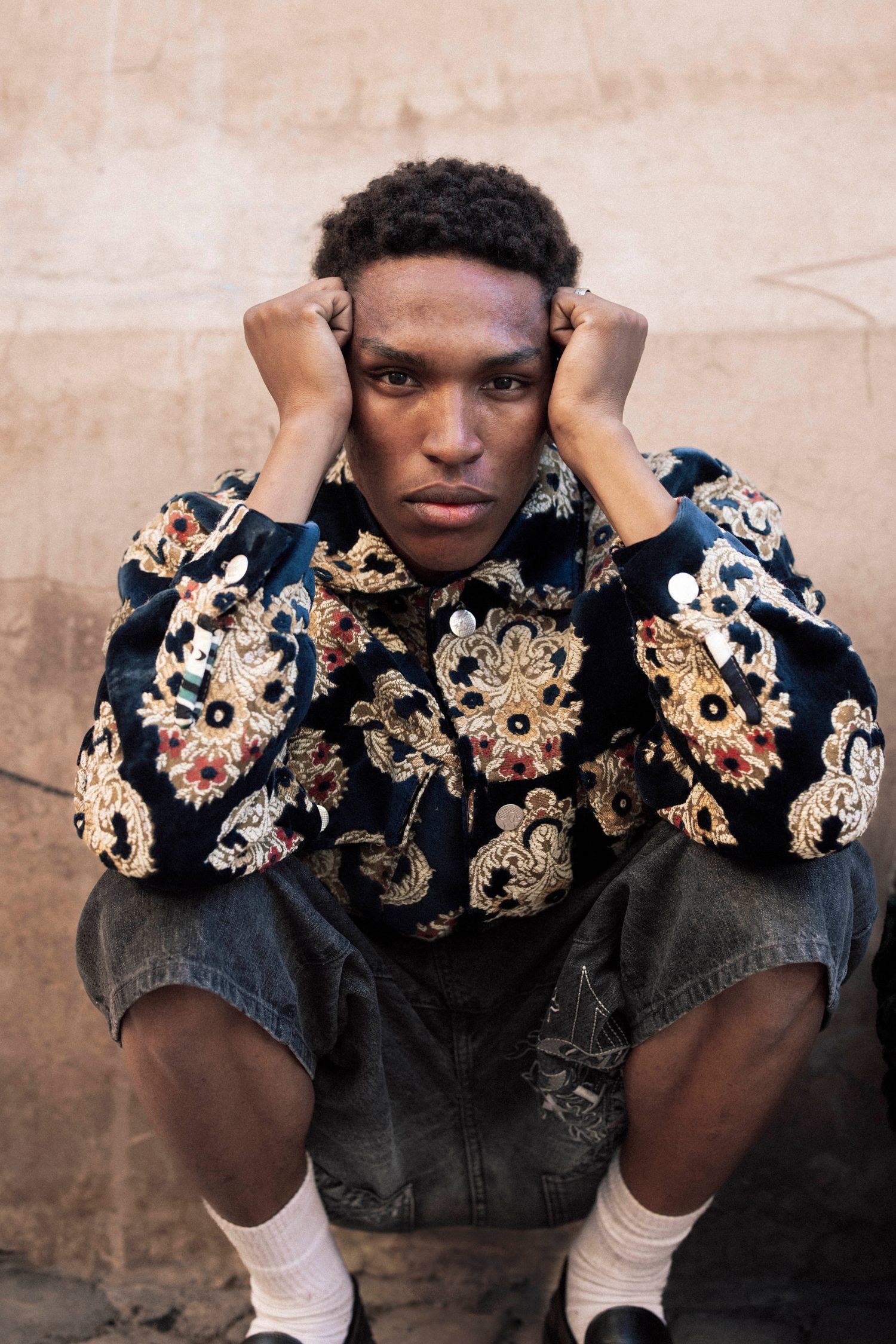 Model in an embroidered velvet jacket crouching with hands on his head