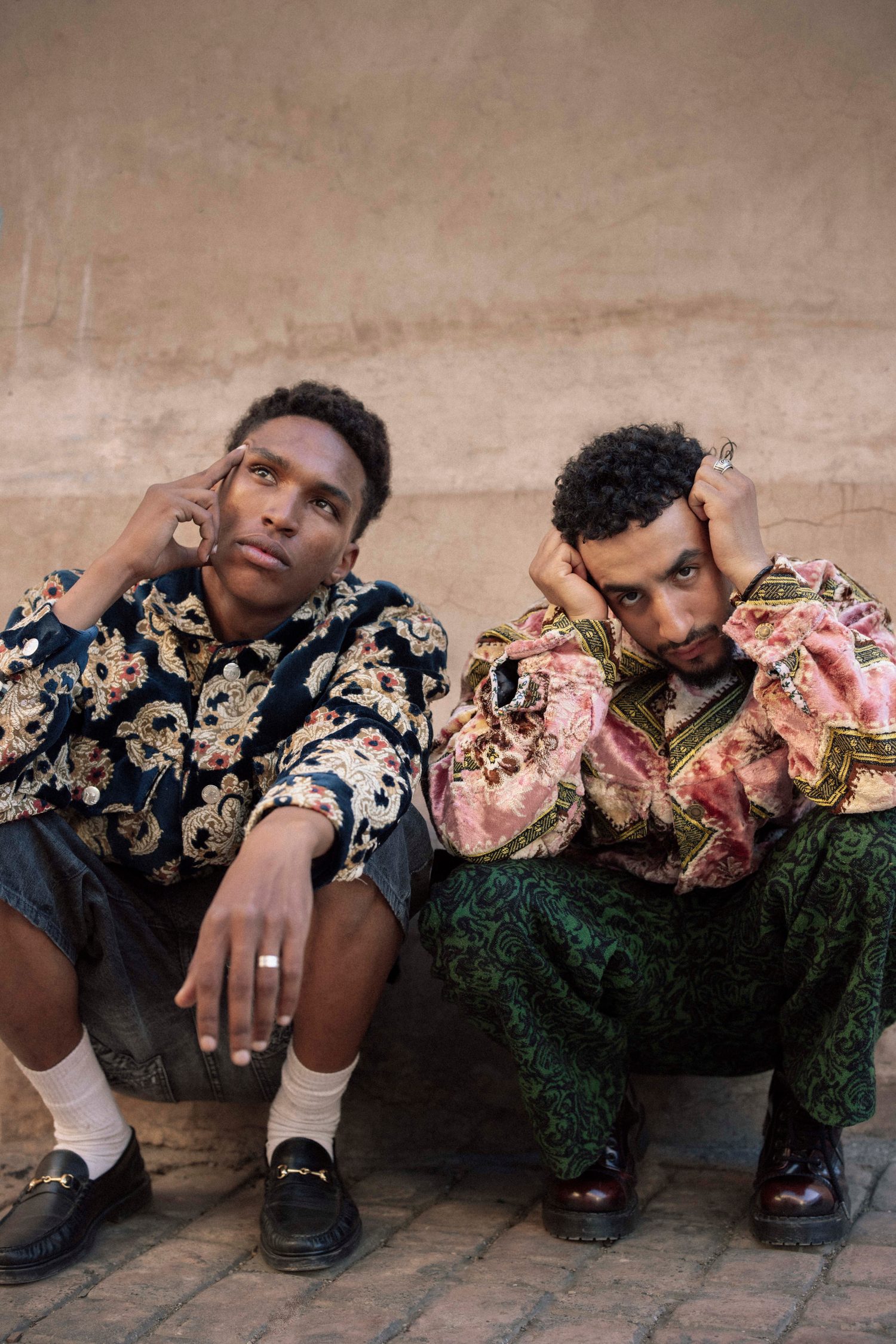 Two models in embroidered jackets crouching against a Marrakech wall, hands to their heads