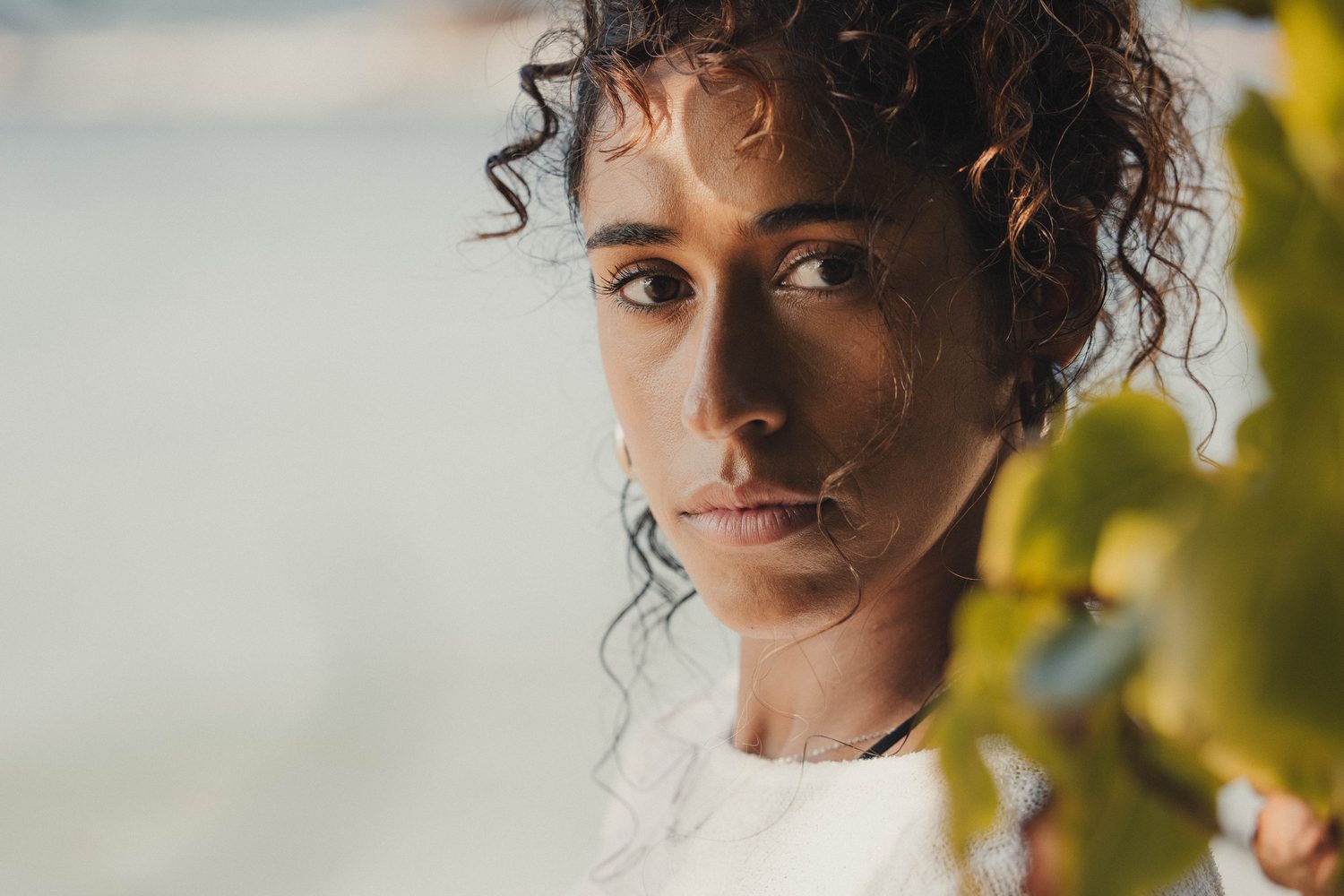 Woman with curly hair in a white knit, framed by leaves on the beach