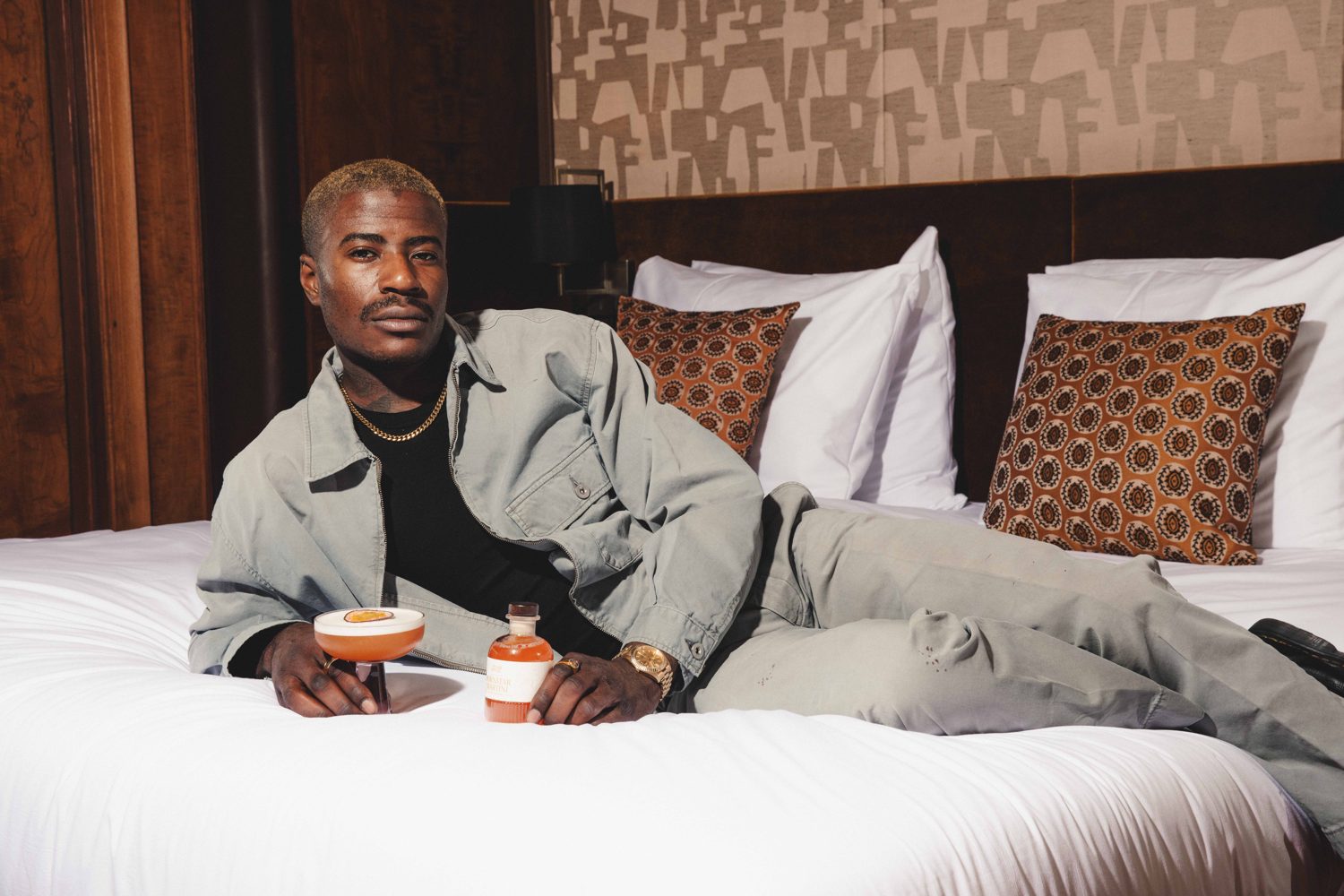 Man in a grey suit lounging on a hotel bed with two cocktails