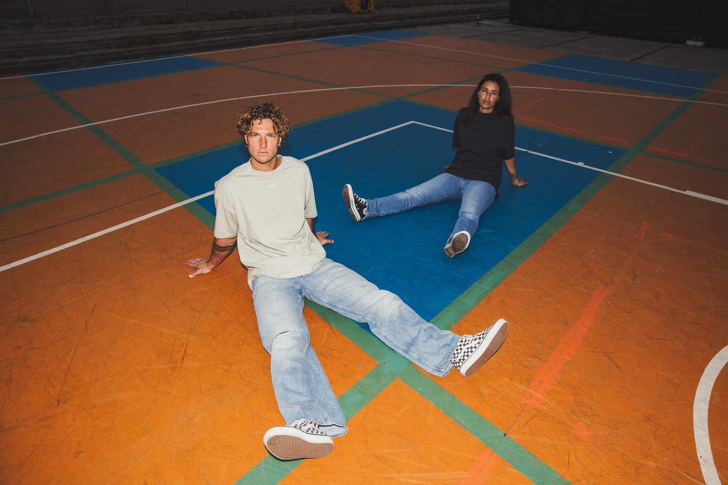 Two models in Buéno T-shirts sitting on an orange and blue basketball court