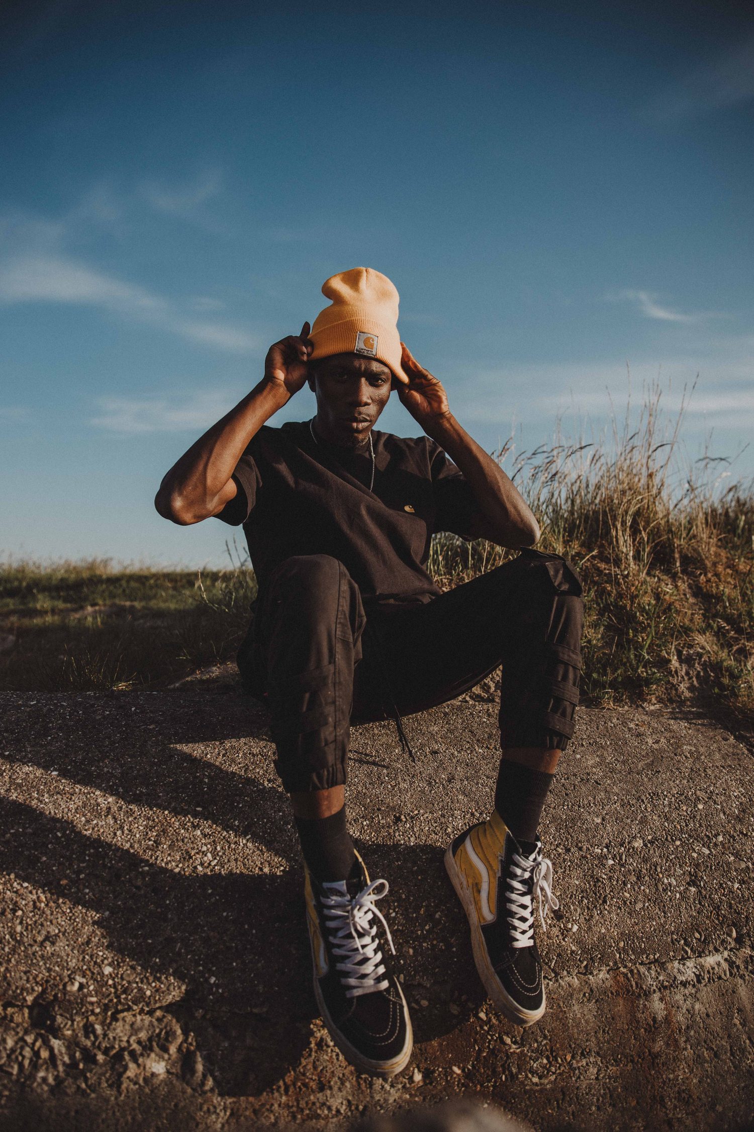 Model in an orange beanie crouching on rocky ground, hands on his head