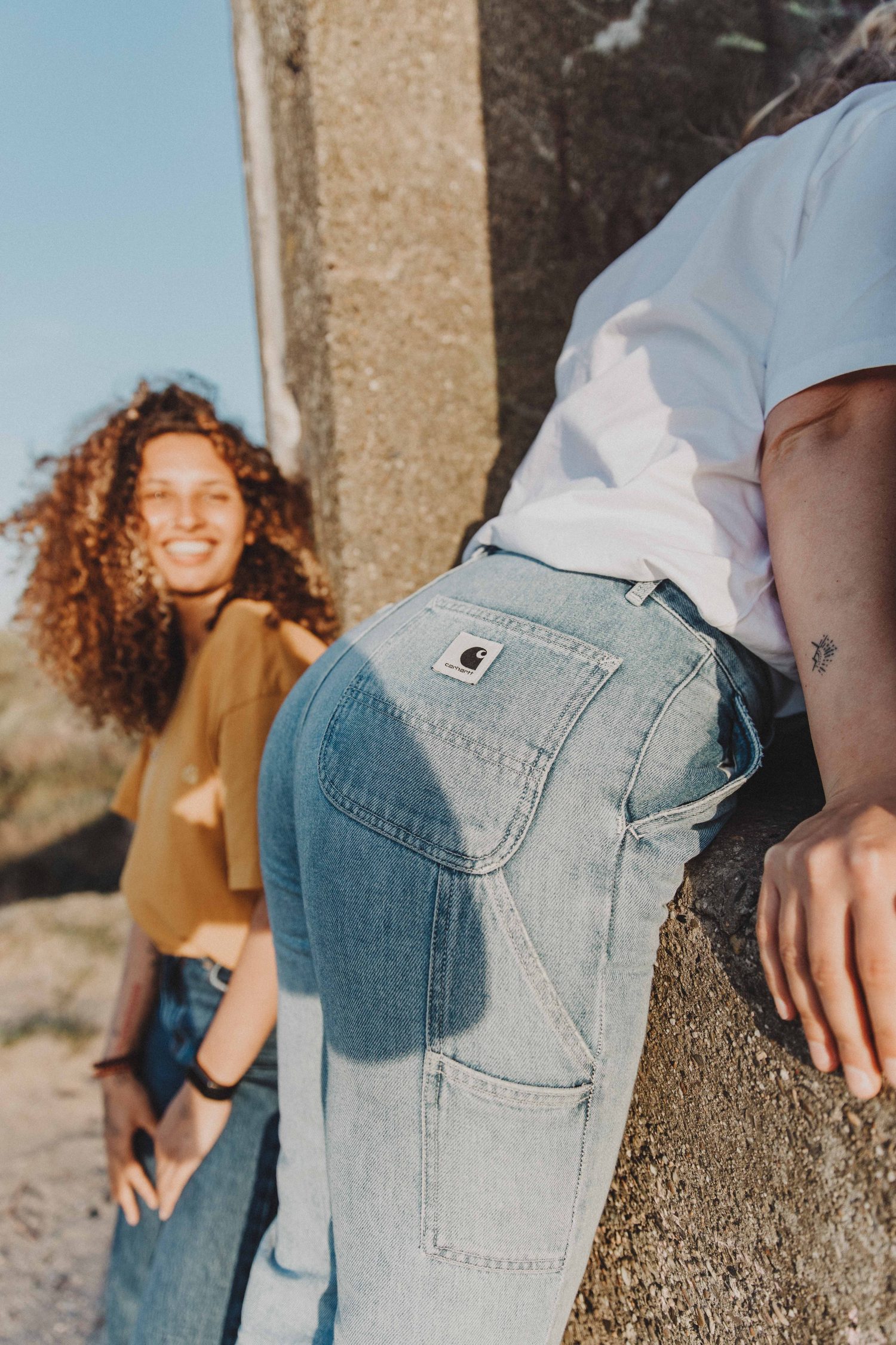 Curly-haired woman smiling behind someone in light jeans with a Carhartt patch