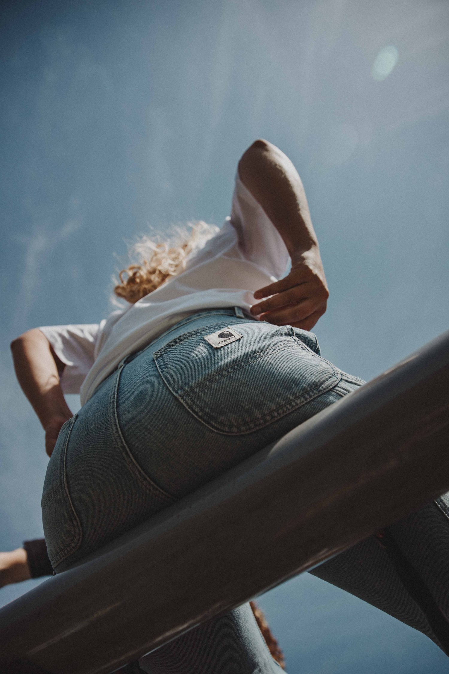 Model in light Carhartt jeans sitting on a railing against the sky, seen from below