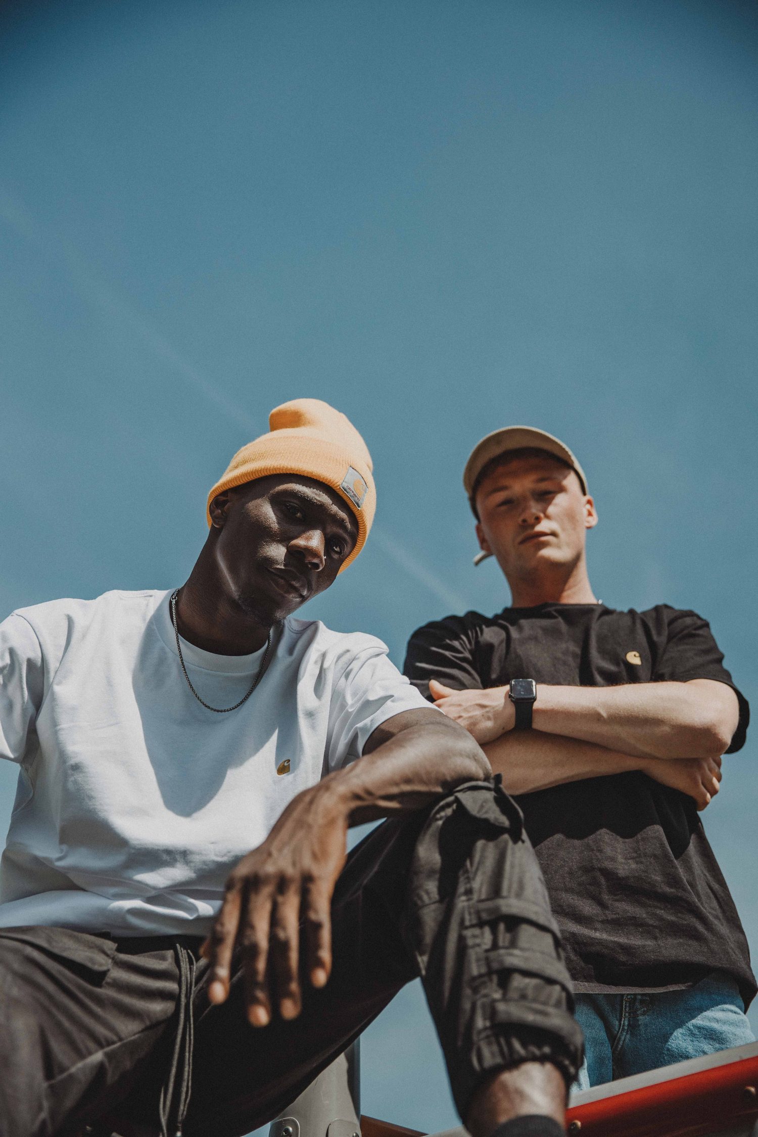 Two models in a beanie and a cap sitting on a climbing frame, arms crossed