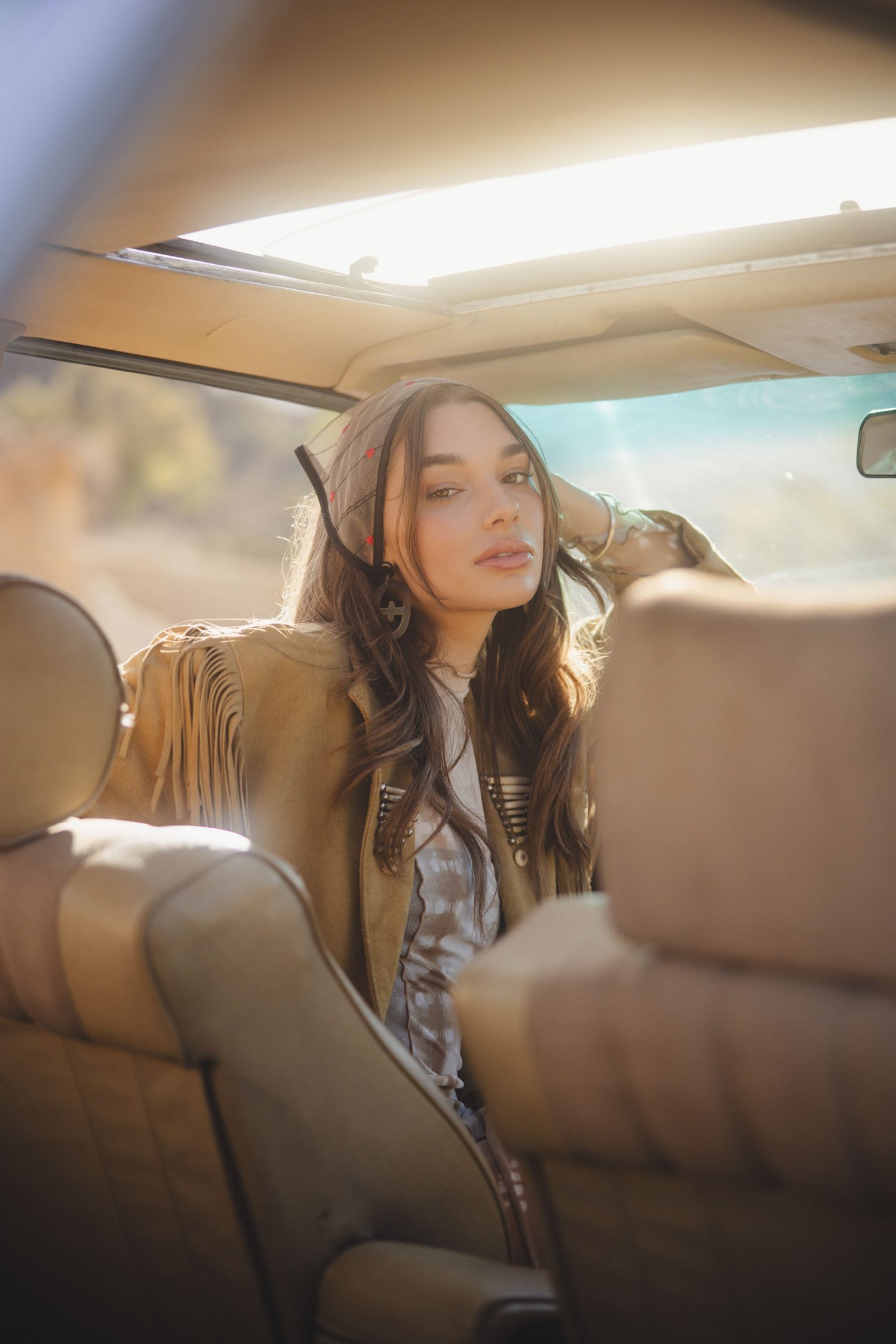 Woman in a fringe jacket on the back seat of a vintage car in warm sunlight