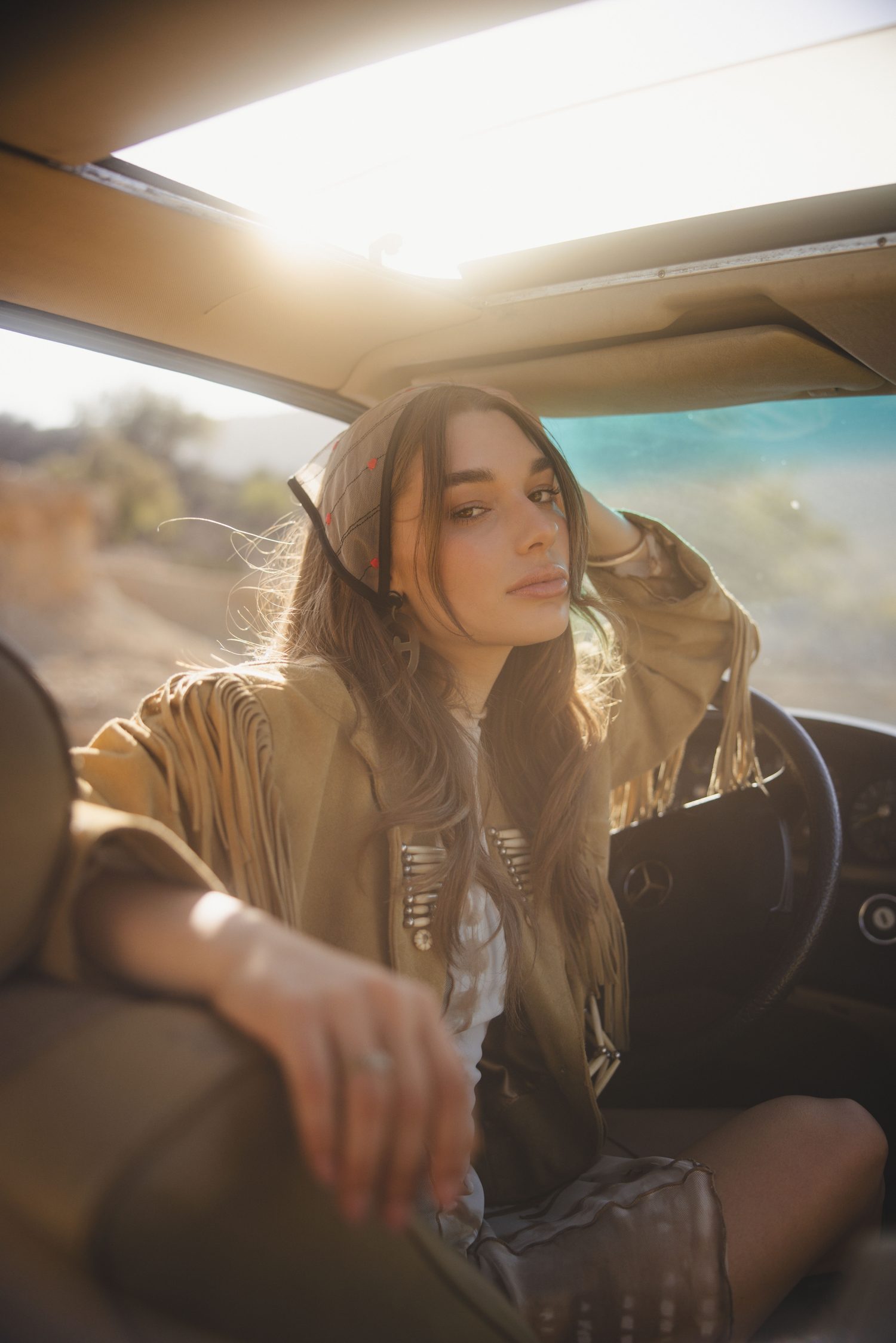 Woman in a suede fringe jacket in the back seat of a vintage car, sun flaring through the roof