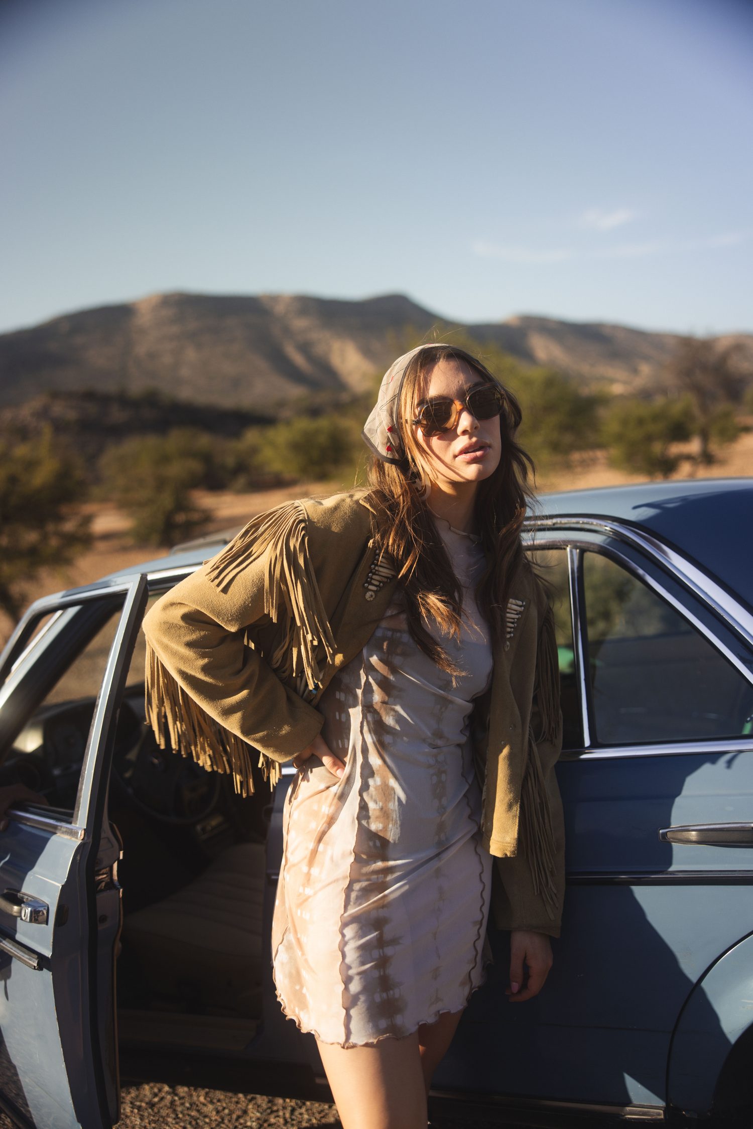Woman in a fringe jacket leaning on the open door of a blue car