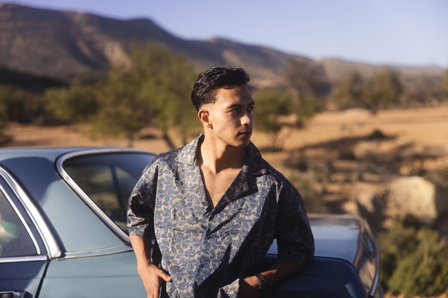 Man in a patterned shirt standing by a vintage car in the mountains
