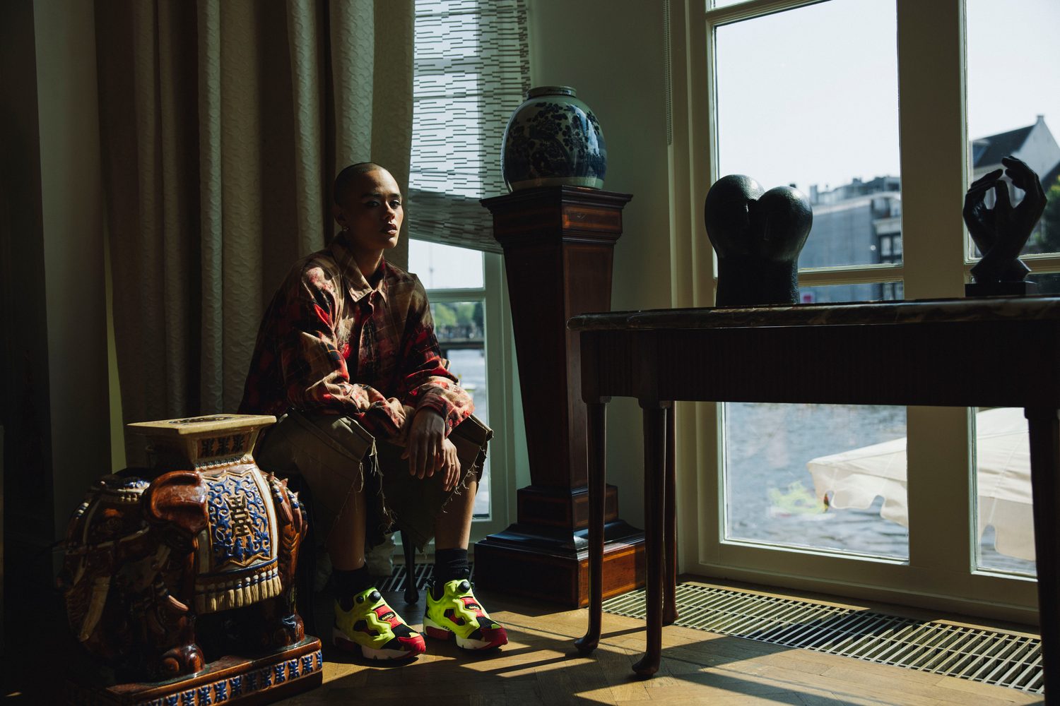 Model in a patterned jacket seated by a sunlit window beside a ceramic elephant stool