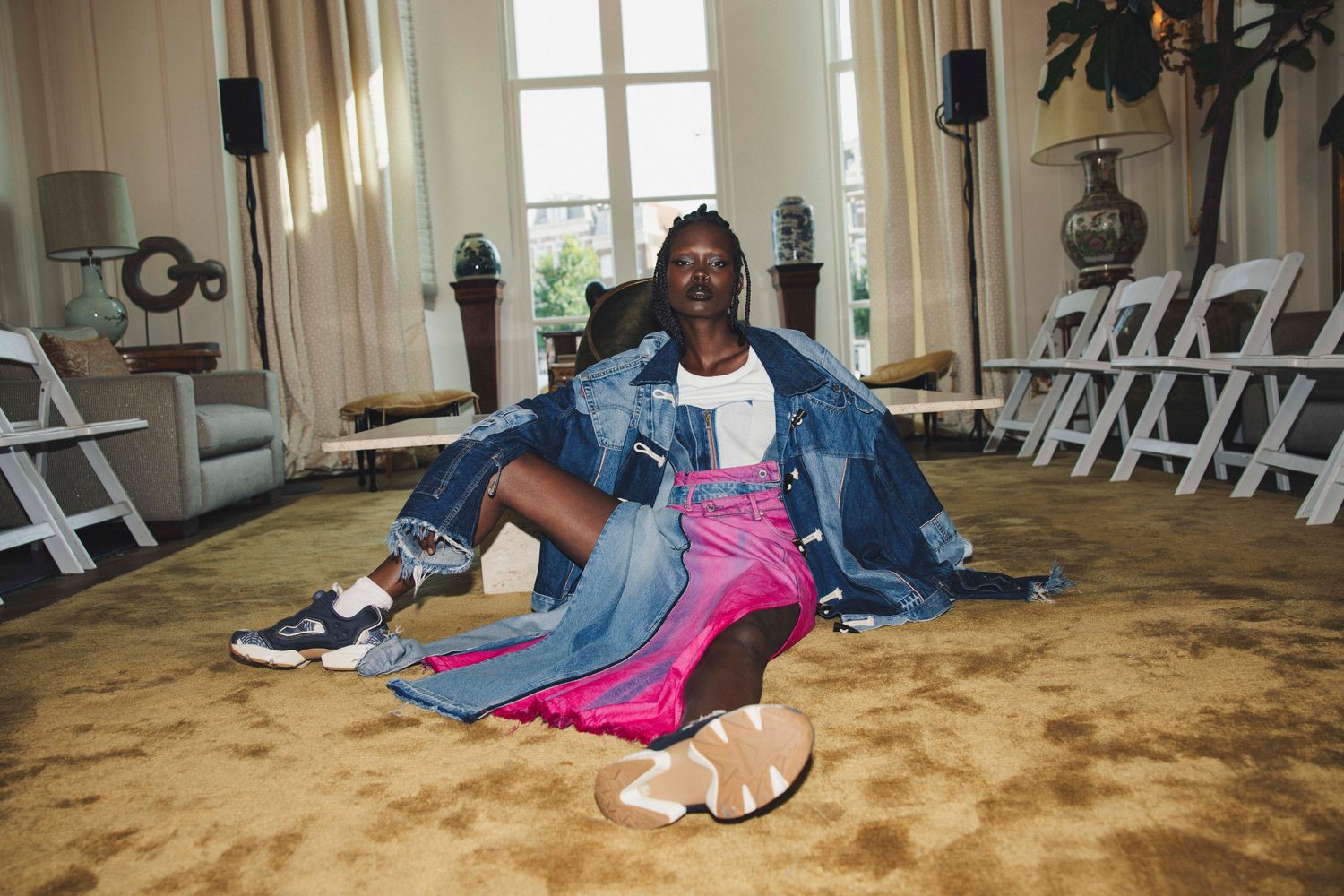 Model in denim and pink seated on the floor of a salon with white chairs