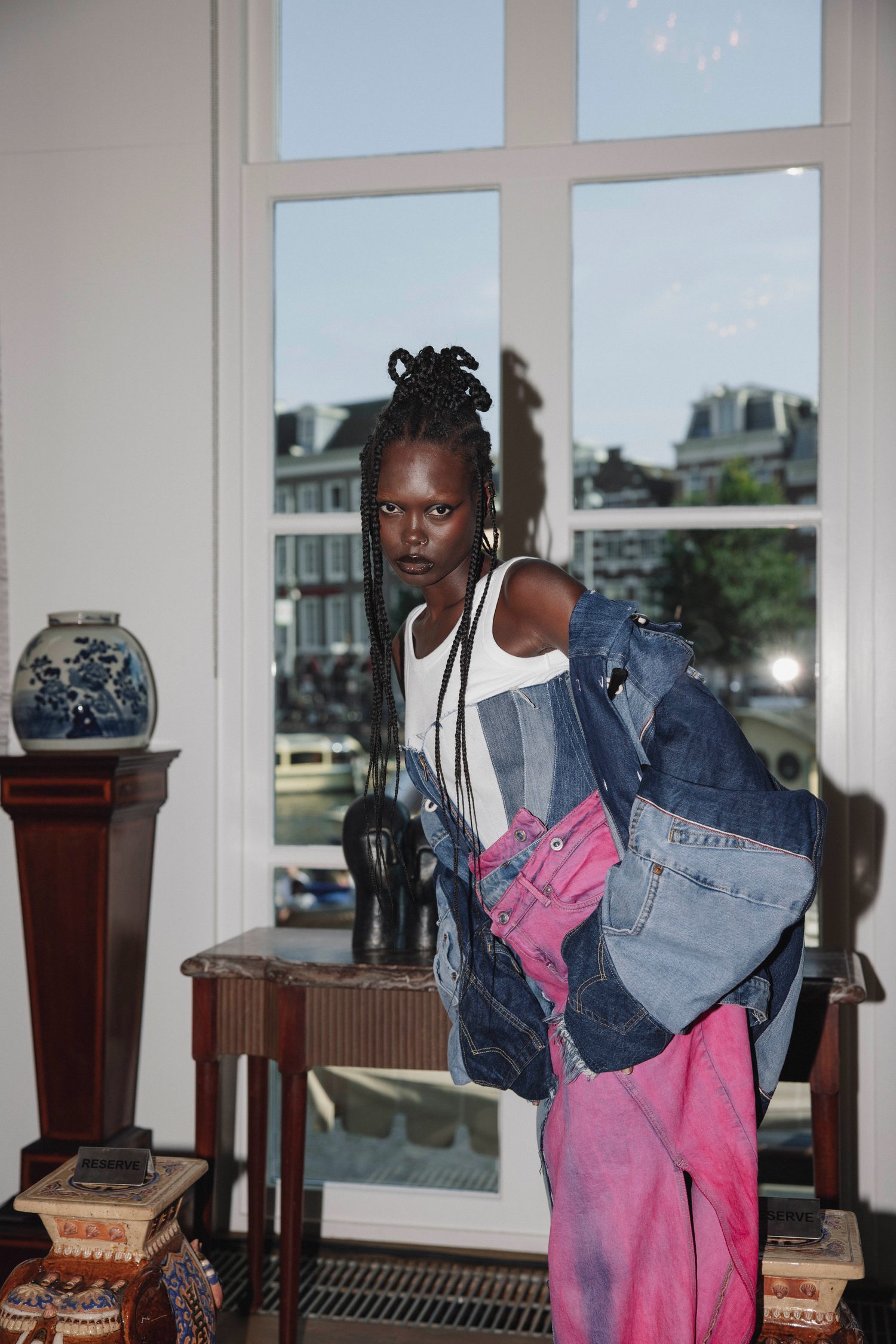 Model with long braids in patchwork denim leaning by a window